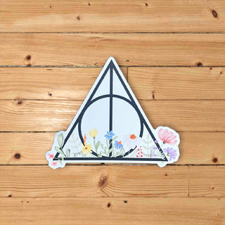 A die-cut sticker featuring the Deathly Hallows symbol from Harry Potter decorated with colorful wildflowers, shown on a light brown wooden plank background.