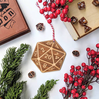 A wooden D20 hanging ornament with engraved numbers and a red-and-white string, styled on a white surface with red berries, evergreen branches, and brown polyhedral dice.