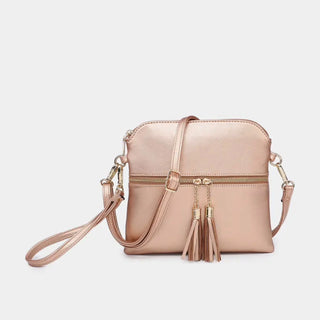 A metallic rose gold crossbody handbag with a zippered front, matching shoulder strap, and decorative tassels, shown against a light gray background.