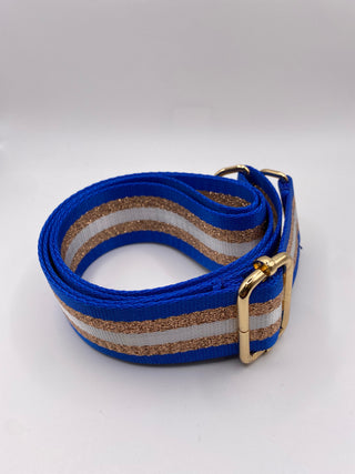 A woven web belt with royal blue, white, and gold glitter horizontal stripes, featuring a gold-tone metal buckle, photographed on a light gray background.