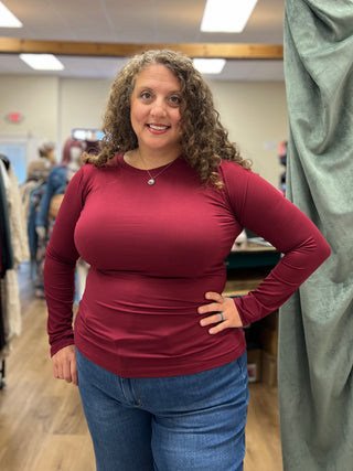 A woman with curly brown hair wearing a fitted, long-sleeve burgundy top and blue jeans, standing in what appears to be a clothing boutique or fitting room.