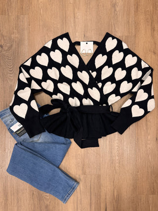 A black knit wrap-style cardigan with a cream heart pattern and a tie waist, laid flat on a wood floor next to a pair of blue jeans.
