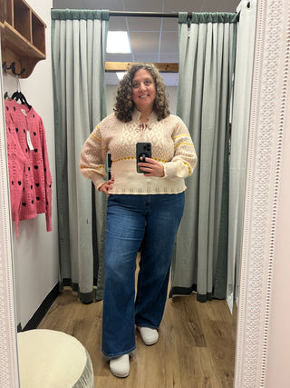 A woman takes a mirror selfie in a fitting room wearing a cream-colored knit sweater with yellow stripes on the sleeves and blue wide-leg jeans.