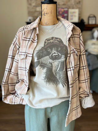 A mannequin torso styled with a beige and brown plaid flannel overshirt layered over a cream graphic T-shirt featuring a black-and-gray dog illustration, paired with light blue pants in an indoor room setting.