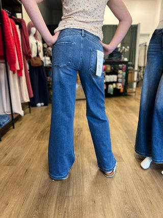 A person standing in a clothing store, photographed from the back while wearing blue wide-leg jeans with a white price tag attached to the back pocket.