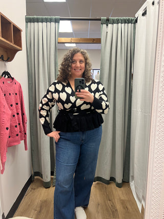 A woman takes a mirror selfie in a fitting room wearing a black and white patterned long-sleeve top with a black ruffled hem, blue jeans, and white sneakers. A pink knit top is hanging on a hanger below her.