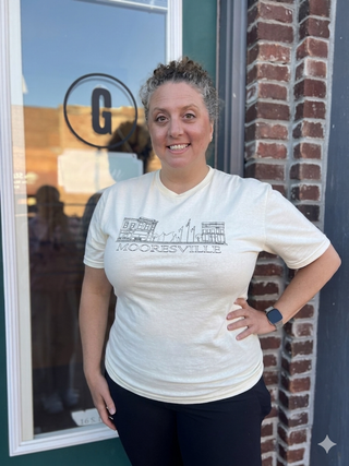 A woman wearing a cream-colored v-neck t-shirt with a black graphic of buildings and the text 'MOORESVILLE' on the front, standing outdoors next to a brick wall and storefront window.