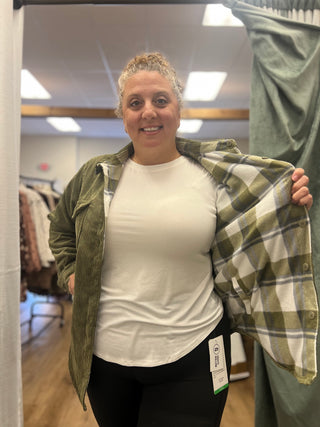 A woman in a fitting room wearing a white t-shirt, black leggings, and holding open an olive green corduroy overshirt with a green, white, and gray plaid lining.