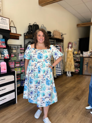 A woman wearing a blue and white floral off-the-shoulder dress stands inside a boutique shop with wooden floors and shelves of accessories.