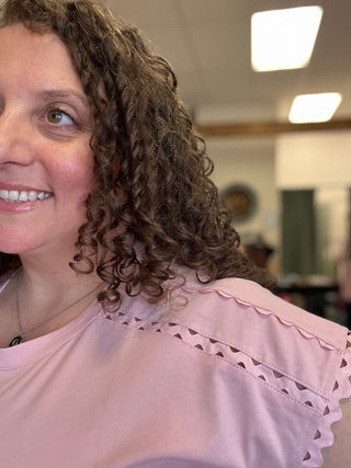 A close-up of a smiling woman with curly brown hair wearing a light pink top with scalloped eyelet detailing on the shoulder.