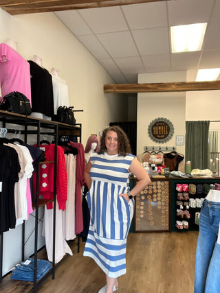 A woman modeling a blue and white striped midi dress inside a clothing boutique, surrounded by racks of folded garments.