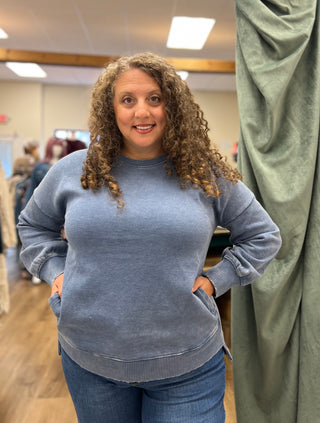 A woman with curly brown hair wearing a heathered slate blue crewneck sweatshirt and blue jeans, standing in a fitting room or boutique.