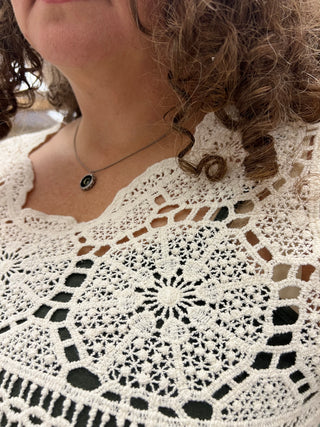 Close-up of a person wearing a white crocheted lace top with an intricate floral and geometric openwork pattern, paired with a silver necklace.