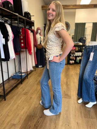A young woman stands in a clothing boutique wearing light-wash blue flare jeans and a cream floral top. Clothing racks and mannequin legs are visible in the background.
