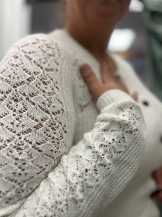 Close-up of a person wearing a cream-colored open-knit sweater with a textured lace pattern on the sleeves and a ribbed crew neck.