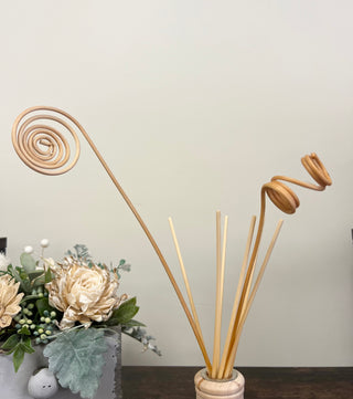 A natural wood reed diffuser with spiral-shaped decorative reeds standing in a round wooden base, displayed on a dark tabletop next to a vase of cream-colored artificial flowers against a plain light wall.