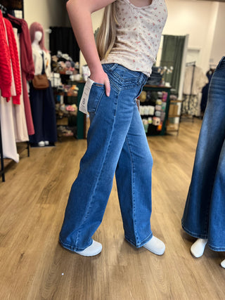 A person standing in a clothing store wearing high-waisted, wide-leg blue jeans with a cream floral tank top. The jeans have a visible white size tag hanging from the waistband and are photographed against a background of wood floors and store fixtures.