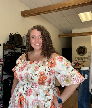 A smiling woman with curly brown hair wearing a white off-the-shoulder dress with a vibrant orange, pink, and green floral print, standing inside what appears to be a boutique.