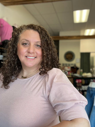 A smiling woman with curly brown hair wearing a light pink ribbed short-sleeve top, photographed in a retail store setting.
