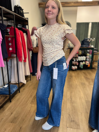 A woman modeling a beige short-sleeve top with a small floral print and blue wide-leg jeans in a clothing store.