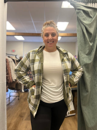 A woman stands in a fitting room wearing a green and white plaid hooded flannel shirt over a white t-shirt and black leggings.