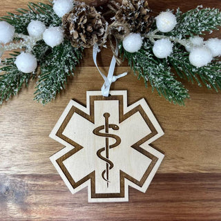 A wooden Star of Life ornament with a laser-engraved medical caduceus symbol, hanging from a white ribbon and styled with a snowy pine branch and pinecones on a wooden surface.