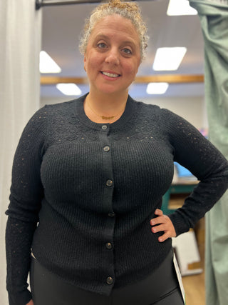 A woman wearing a dark charcoal gray lace-trimmed button-up cardigan sweater, standing in what appears to be a fitting room or boutique.
