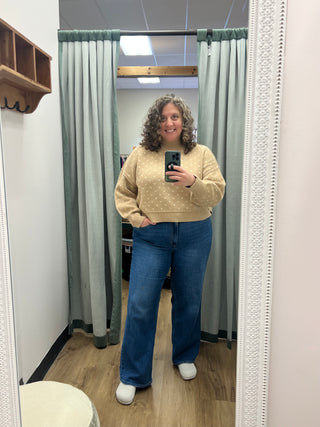 A woman takes a mirror selfie in a fitting room wearing a beige long-sleeve knit sweater with a subtle white polka dot pattern and blue wide-leg jeans.