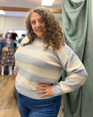 A woman with curly brown hair wearing a waffle-knit sweater with horizontal stripes in cream, light blue, and gray, paired with blue jeans, standing in a fitting room.