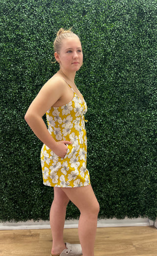 A woman modeling a yellow and white floral print mini dress, standing in front of a green hedge wall.