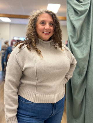 A woman with curly brown hair wearing a beige ribbed mock-neck knit sweater and blue jeans, standing in an indoor boutique setting.