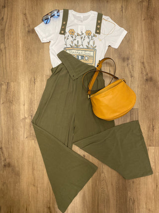 A styled flat-lay outfit on a wood floor featuring olive green wide-leg pants, a white graphic t-shirt with floral suspenders, a mustard yellow crescent-shaped leather bag, and blue-tinted sunglasses.