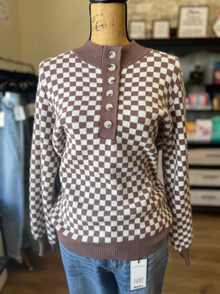 A brown and white checkered knit sweater with a buttoned henley collar, displayed on a mannequin torso with blue jeans in a retail store setting.