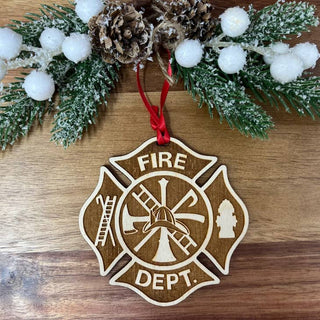A wooden Christmas ornament shaped like a Maltese cross with a fire department emblem, hanging from a red ribbon on a wooden surface with holiday greenery and white pom-poms above it.