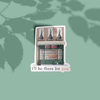 A die-cut sticker depicting a quaint brick storefront with a striped awning, a bicycle, and the text 'I'll be there for you.' in black and peach lettering, shown on a muted sage green background with soft leaf shadows.