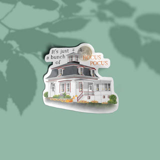 A die-cut sticker featuring an illustration of a white house with a dark gray roof and the text 'It's just a bunch of HOCUS POCUS', shown on a muted green background with soft leaf shadows.