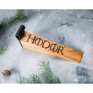 A wooden door stopper engraved with the word 'HODOR' in a gothic font, featuring a black leather loop at one end, displayed on a textured gray surface with pine branches and a small pinecone.