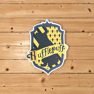 A die-cut sticker of the Hufflepuff crest from Harry Potter, featuring a yellow and navy blue shield with a Hufflepuff puggle and the word 'Hufflepuff' on a banner, displayed on a light brown wooden plank background.