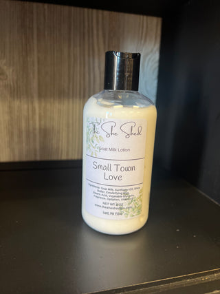 A bottle of The She Shed Goat Milk Lotion labeled 'Small Town Love' sitting on a dark shelf against a wood-paneled background.