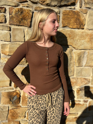 A woman wearing a brown long-sleeve henley top and leopard print pants, standing in front of a stone wall.
