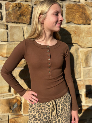 A young blonde woman wearing a brown long-sleeve henley top and leopard print pants, standing outdoors against a stone wall.
