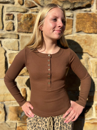 A young blonde woman wearing a brown long-sleeve henley shirt with a button placket, standing in front of a stone wall.