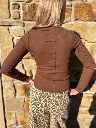 A woman shown from the back wearing a fitted brown long-sleeve top and leopard-print pants, standing in front of a stone wall.