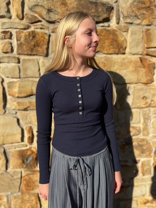 A young blonde girl wearing a navy blue long-sleeve henley top and a gray pleated skirt, standing in front of a rustic stone wall.