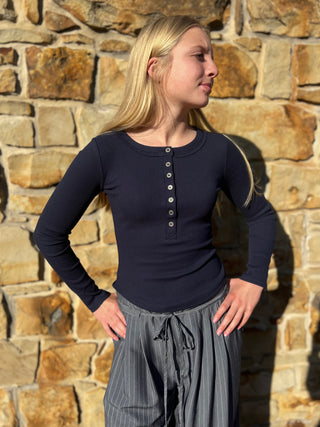 A woman with blonde hair stands in front of a rustic stone wall, wearing a dark navy long-sleeve henley top with a button placket and gray pinstriped pants.