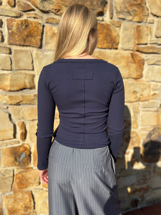 A woman viewed from behind wearing a navy blue long-sleeve ribbed top and gray pinstriped pants, standing in front of a sunlit stone wall.