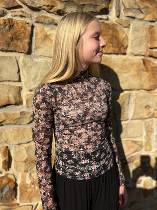 A blonde woman in profile wearing a long-sleeve black mock-neck top with a pink and beige floral pattern, standing against a rustic stone wall.