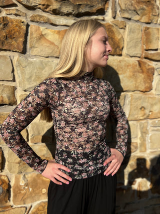 A woman wearing a sheer black long-sleeve mock-neck top with a pink and green floral pattern, posed against a rustic stone wall.