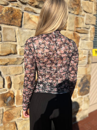 A woman viewed from behind wearing a sheer long-sleeve floral top with a black base and pink flowers, paired with black pants, standing in front of a stone wall.