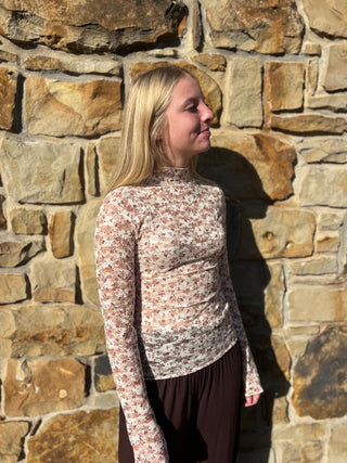 A woman with blonde hair stands in profile against a rustic stone wall, wearing a fitted long-sleeve mock-neck top with a small floral print in beige, brown, and muted red tones, paired with dark brown bottoms.